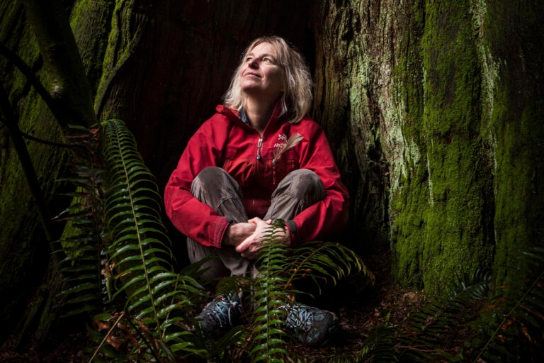 A photograph of Suzanne Simard sitting at the based of a tree