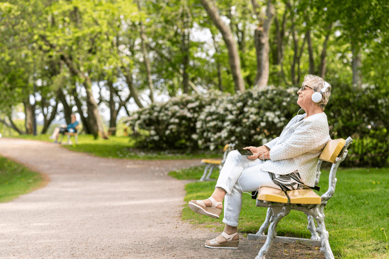 A woman sits on a bench in a park, smiling as she listens to an audiobook