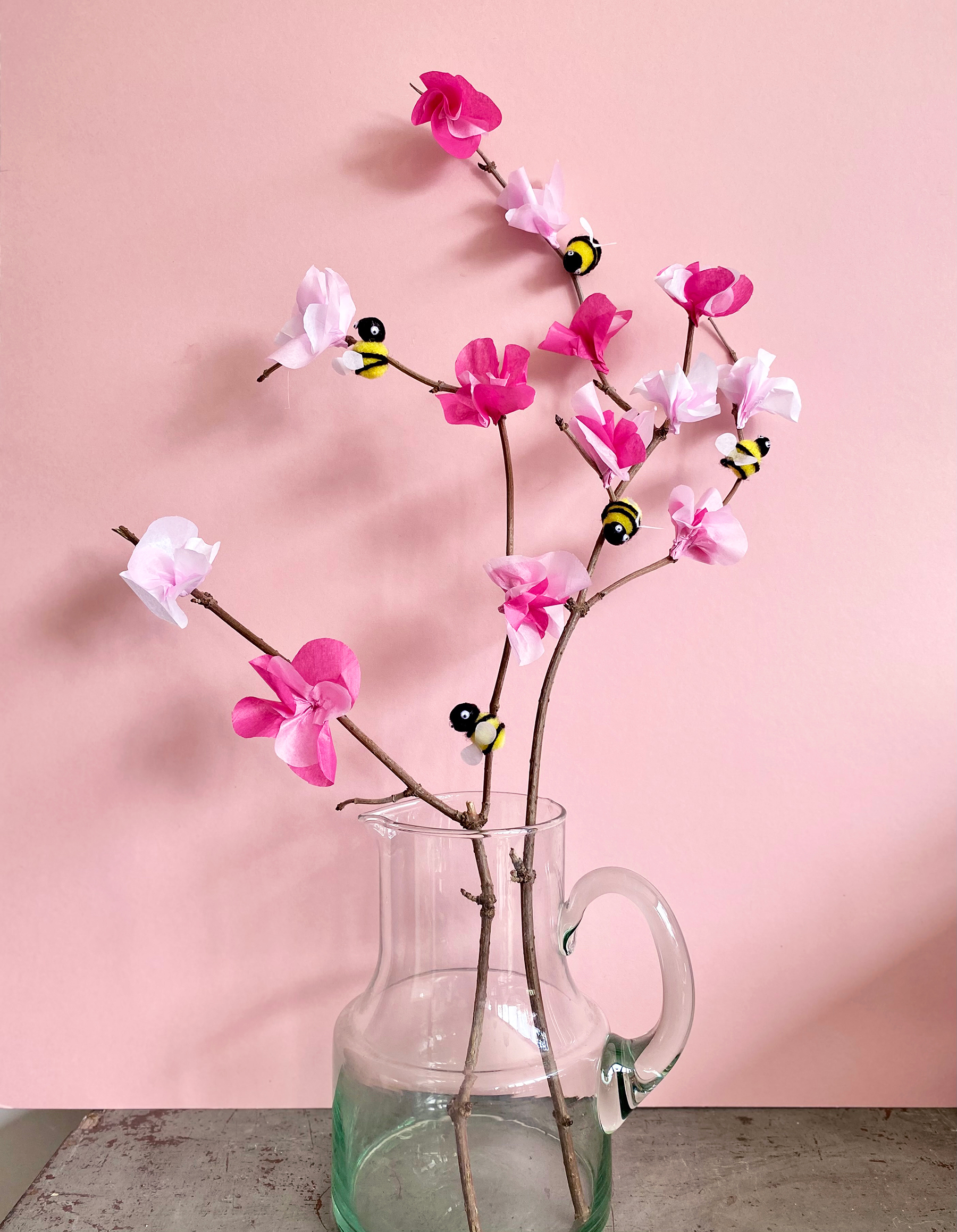 Photo of homemade cherry blossom with pom pom bees in a clear jug against a pale pink wall
