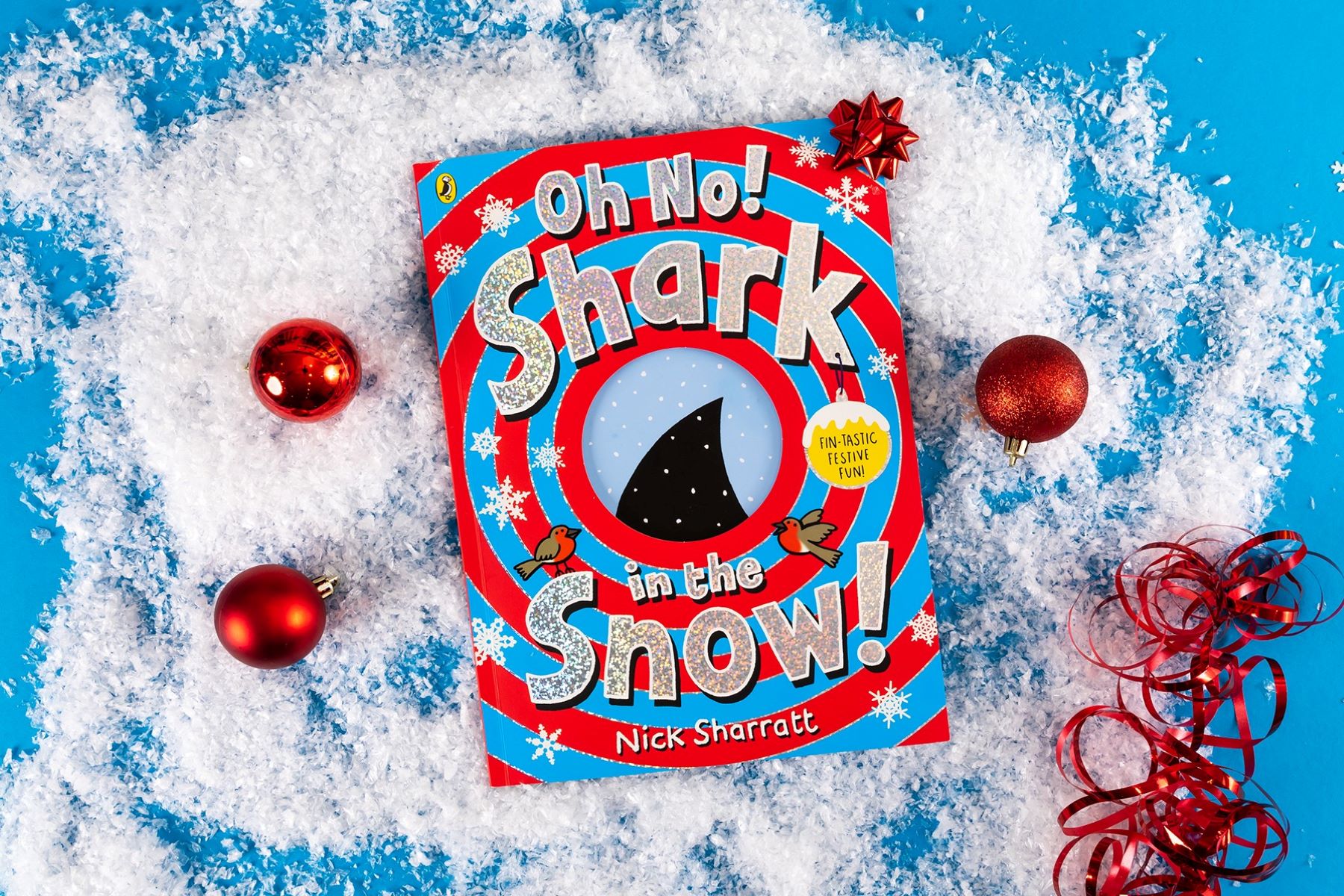 A photo of the book Oh No! Shark in the Snow! on a light blue background surrounded by fake snow and red baubles