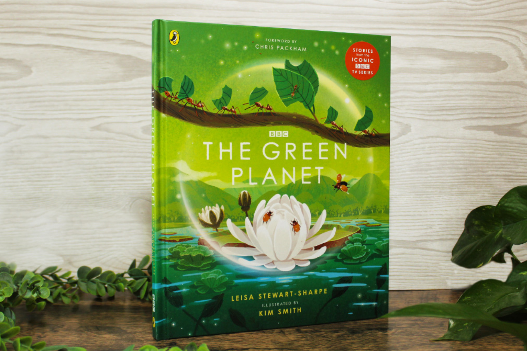 A photo of a copy of The Green Planet book propped up against a wooden background surrounded by plants
